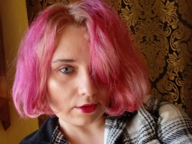JanetParadise with pink hair, blue eyes, red lipstick, and a black-and-white checkered shirt, looking at the camera. Her hair is slightly messy, covering one eye.