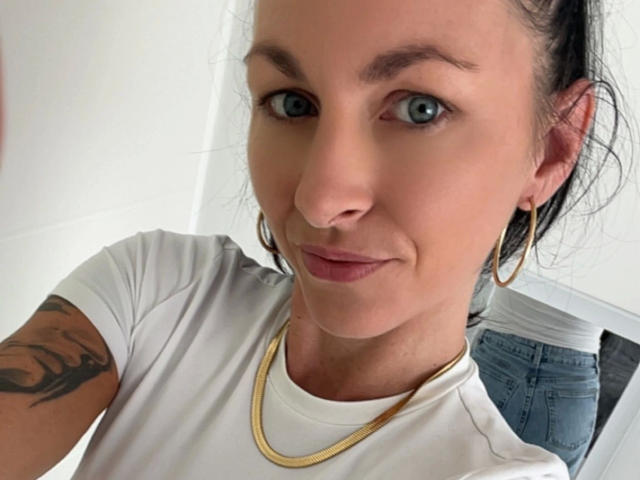 13-Mel-hot with blue eyes, white tee, gold necklace, hoop earrings, and arm tattoo, showing off her ass in blue jeans, smiling slightly.