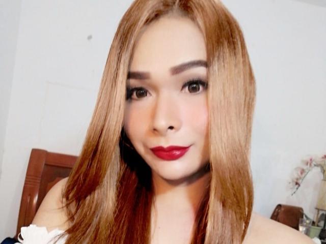 SexyTemptaciousX with long, straight, brown hair, red lipstick, and dark eye makeup, smiling gently at the camera. Her bare shoulders and white top are visible.