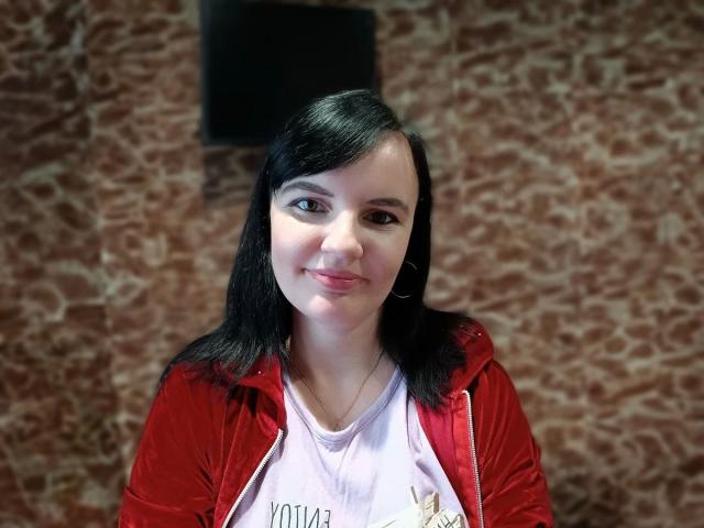 Alicexbaby69-hot with black hair, red velvet jacket, and pink shirt, smiling slightly, hoop earrings, fair skin, brown eyes, medium-sized tits, standing against brown patterned wall.