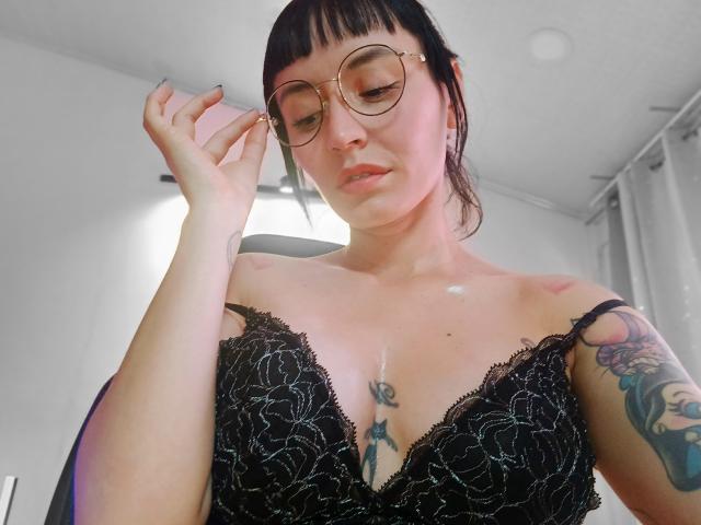SaraleePoison, avec ses lunettes rondes, son soutien-gorge en dentelle noire et ses tatouages, ajuste ses lunettes, montrant ses seins et un petit tatouage sous sa poitrine, dans une pièce très éclairée..