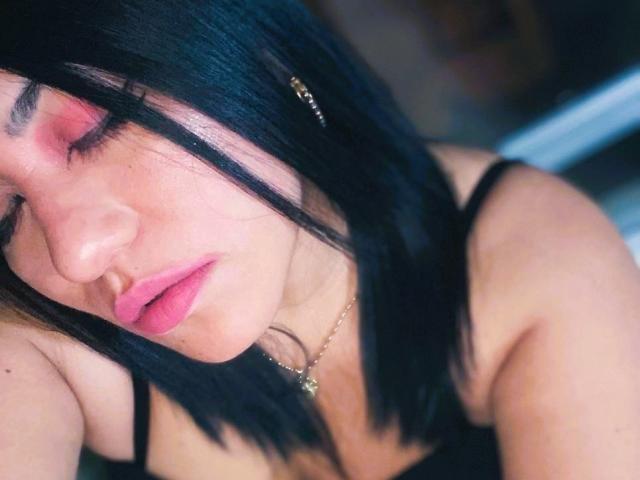 AmaiaCruz with black hair, pink eyeshadow, and pink lips, sleeping with eyes closed, wearing a black spaghetti-strap top, and a gold necklace, looking relaxed.
