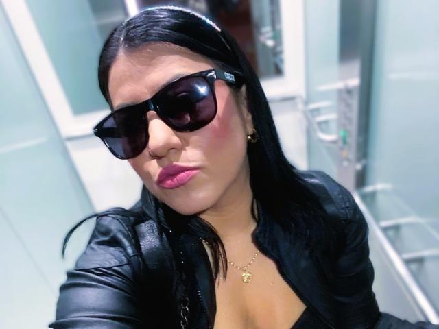 AmaiaCruz with black sunglasses, pink lipstick, and a black leather jacket, showing her cleavage and wearing a gold necklace, has long black hair and a pouty expression.