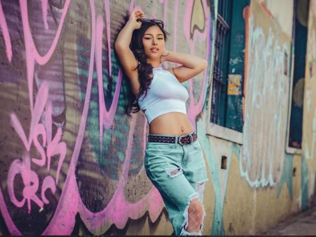 EmilyVv leaning against a graffiti wall, wearing a white crop top, ripped jeans, black belt, and purple sunglasses, with her hands in her hair, showing her small ass.