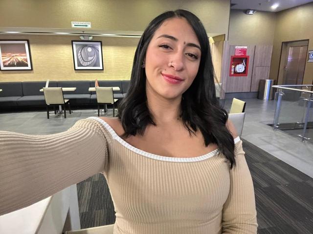 ZaraBeicker with medium tits, beige ribbed top, black wavy hair, pink lipstick, smiling, holding phone, in office or cafe.