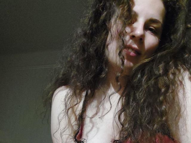 AvaLu, com cabelos castanhos cacheados, sorrindo levemente, vestindo um sutiã vermelho, mostrando seus seios, e olhando para baixo para a câmera. Sua pele é clara, com algumas sardas..
