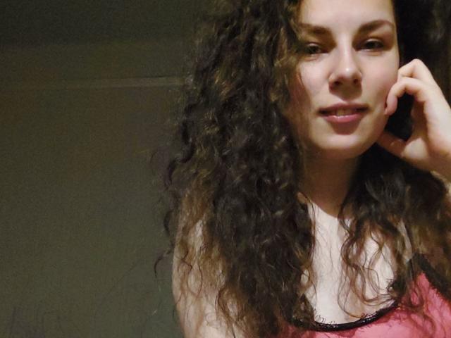 AvaLu, com cabelo castanho encaracolado, blusa rosa com detalhes em renda preta, segurando o telefone no ouvido, sorrindo levemente, com seios médios visíveis, pele clara e expressão relaxada em um quarto com pouca luz..