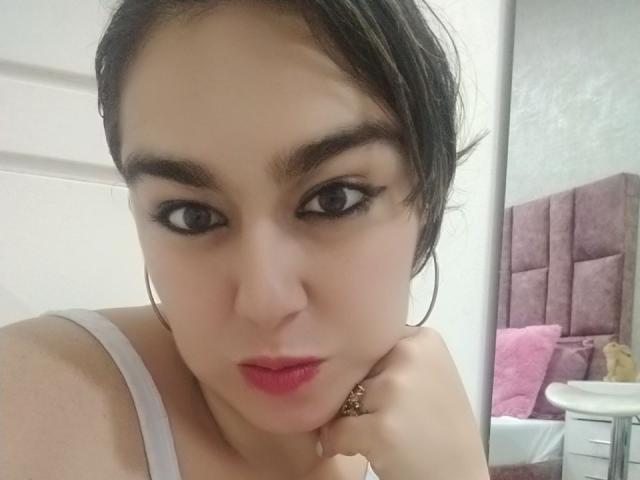 TaylorNova with pouty lips, dark eyebrows, and pink lipstick, wearing a white top, has black hair in a messy bun, and large hoop earrings, looking directly at the camera.