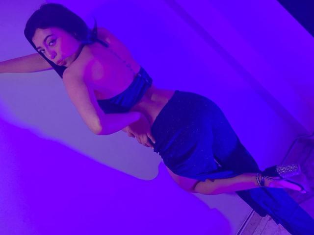SaraHall with dark hair, wearing a black strapless top and black skirt, looking back, hand on hip, purple lighting, high heels, tattoo on right side of her waist.