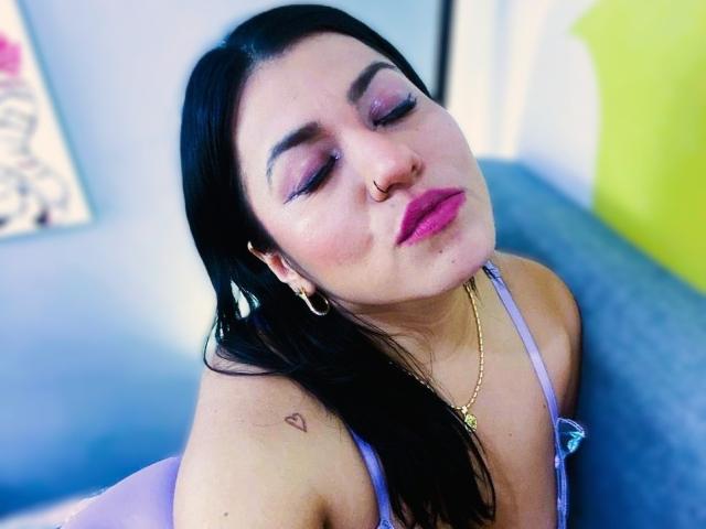 AmaiaCruz with black hair, purple eyelid makeup, pink lipstick, nose ring, hoop earrings, gold necklace, small heart tattoo on shoulder, wearing lilac top, eyes closed, slight pout.