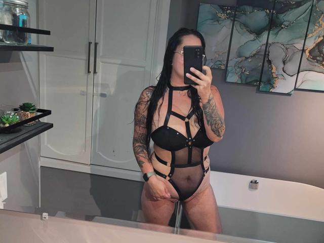 RoriQ stands in a bathroom wearing black strappy lingerie, exposing her tits and pussy. She has tattoos on her arm and is taking a mirror selfie with her phone.