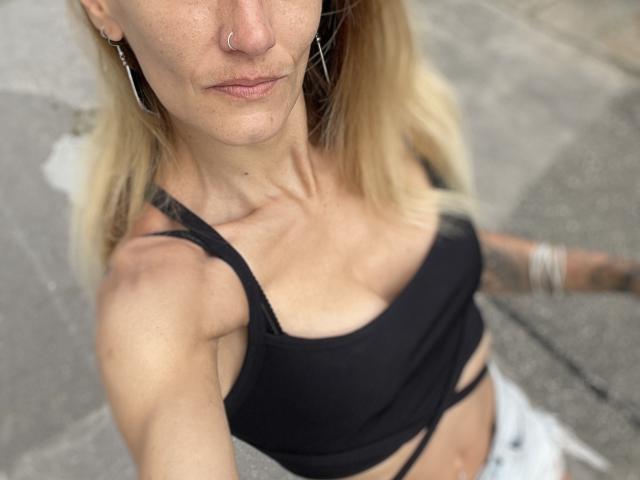 DaneeSoHot with blonde hair, nose ring, hoop earrings, black crop top showing small tits, tattooed arm, and white jeans, taken from a high angle.