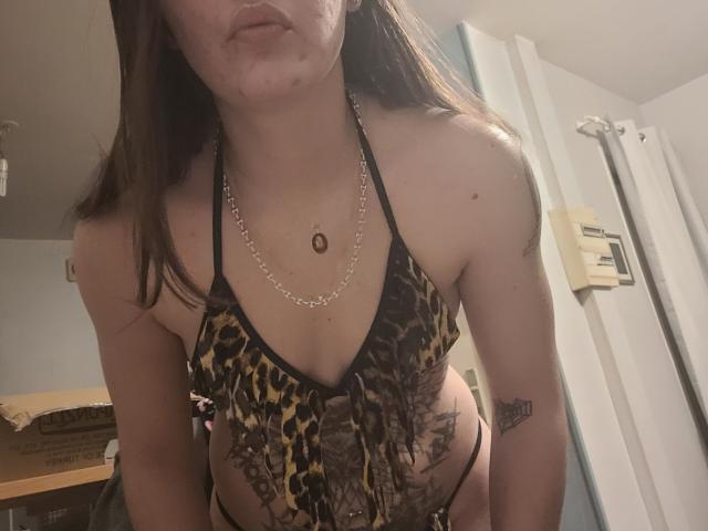 MiaGia, com cabelo castanho, lábios carnudos, vestindo uma blusa de alça com estampa de leopardo, expondo seus seios e um tatuagem no braço direito, e um colar de corrente..