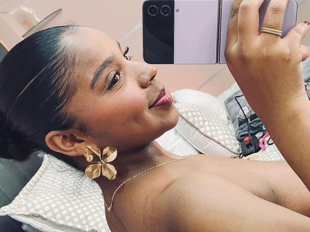 LisaMendez, com pele escura, pequenos brincos de borboleta dourados e batom vermelho, tirando uma selfie com um telefone rosa, mostrando seus ombros nus e um leve sorriso..