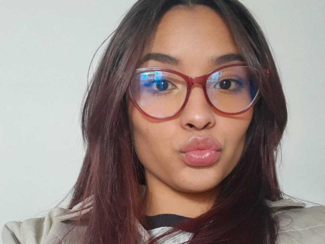 LeaKlarck has dark brown hair, red glasses with a blue reflection, and is pouting her shiny pink lips. She's wearing a light gray jacket with a black top.