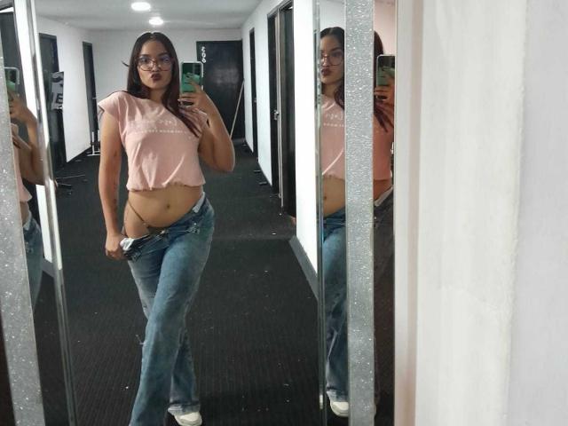LeaKlarck with glasses, pink crop top, blue jeans, and white sneakers, showing her bare lower belly and pulling down jeans slightly, standing in a mirror selfie.
