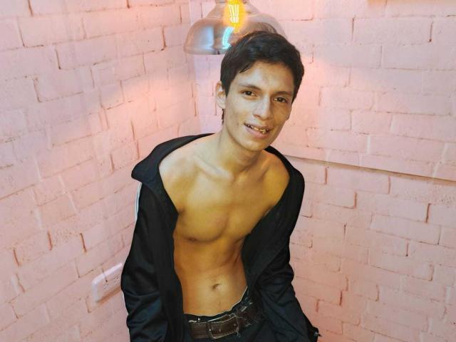 BastianClark, shirtless with a black hoodie, has a slim, toned, brown-skinned body, short dark hair, and a slight smile, standing in a corner with a hanging light.