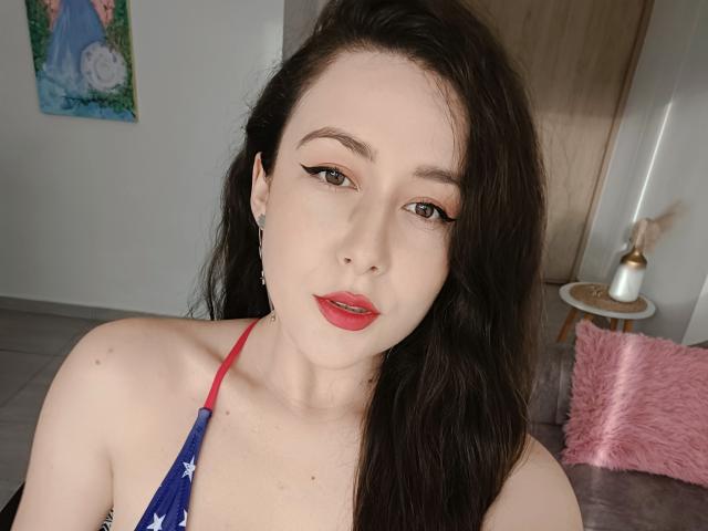 MabyLegrand with pale skin, long dark hair, red lipstick, eyeliner, wearing a halter top with a blue American flag pattern, showing her bare shoulders and small tits.