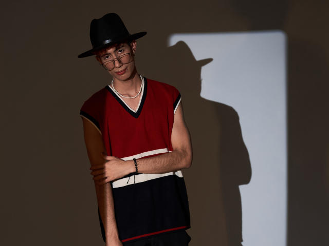 NovaWheeler in a black hat, red sleeveless top, and black pants, with glasses and a bracelet, standing with arms crossed, shadow on the wall.