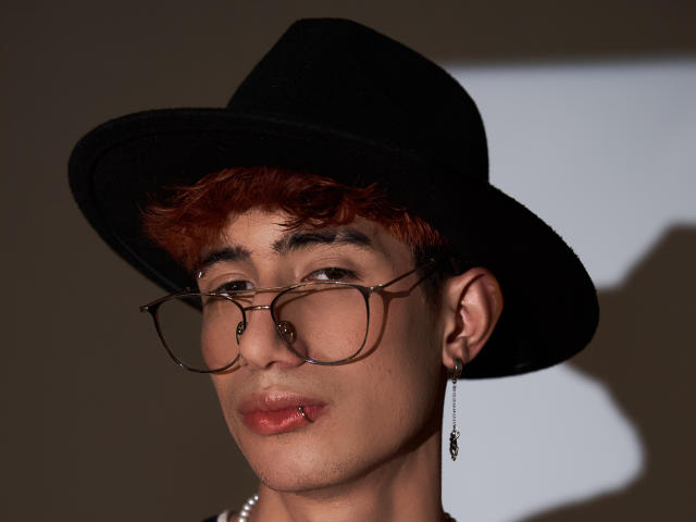 NovaWheeler with red curly hair, black hat, brown glasses, nose ring, and earring, looking slightly to the side with a subtle pout. Soft shadow on the right.