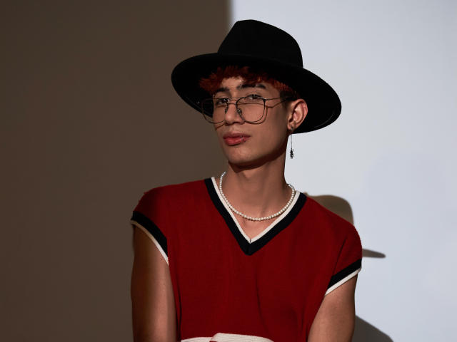 NovaWheeler with curly red hair, black hat, glasses, pearl necklace, red V-neck shirt with black trim, subtle smile, shadow on white wall, standing, looking slightly to the left.