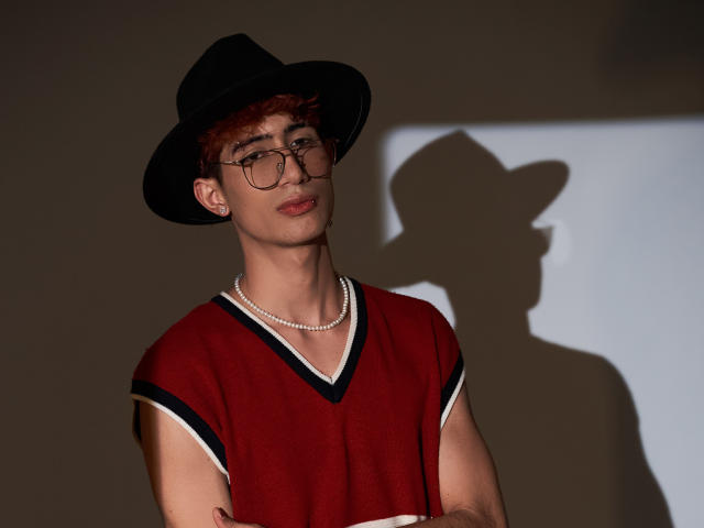 NovaWheeler with red hair, glasses, black hat, pearl necklace, red V-neck shirt, shadow on wall. Hands crossed, slightly smiling. Slim, cute, stylish.
