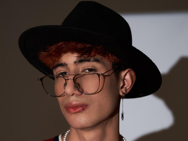 NovaWheeler with curly red hair, black hat, glasses, and earring, pouty lips, shadow on face, wearing pearl necklace, brown eyes, looking slightly to the side, serious expression.