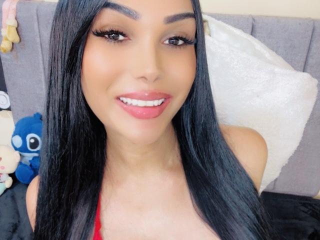 VanezzTss has long black hair, brown eyes, and glossy pink lips. She's wearing a red top, showing her small tits. Her makeup includes long eyelashes.