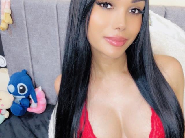 VanezzaTss with long black hair, wearing a red lace bra, showing her big tits, sitting on a bed with stuffed toys, smiling at the camera.