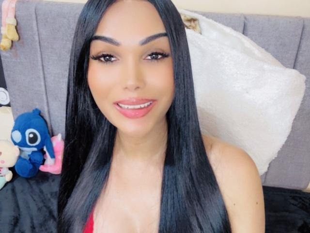 VanezzaTss with long black hair, red lipstick, and large tits in a red bra, smiling at the camera. She's holding her tits with one hand.