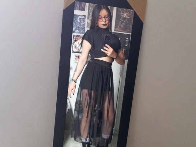 EvangelineJones with black lipstick, glasses, and a sheer black dress, showing her pussy through the fabric, standing in front of a mirror, holding a phone.