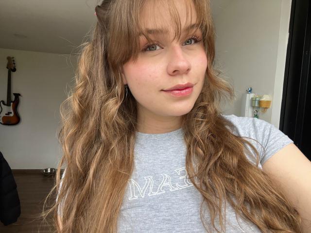 SwanKyivy has wavy, light brown hair, green eyes, and a slight smile. She's wearing a gray t-shirt, showing her small tits, and has pink lipstick on.
