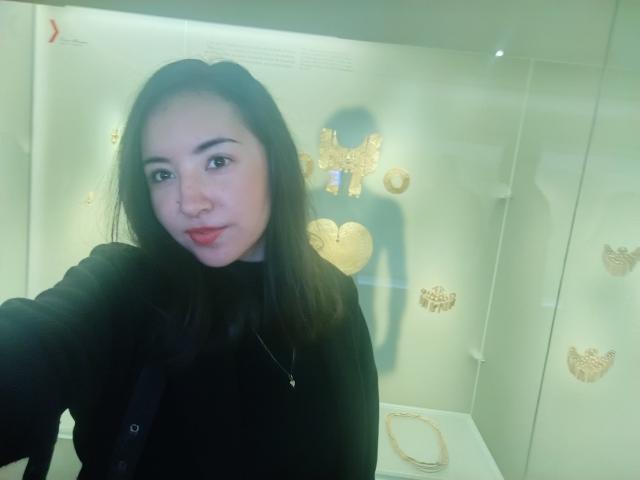 SusanMonroy with black hair, light skin, and red lipstick, wearing a black top, taking a selfie in a museum display with gold jewelry in the background.