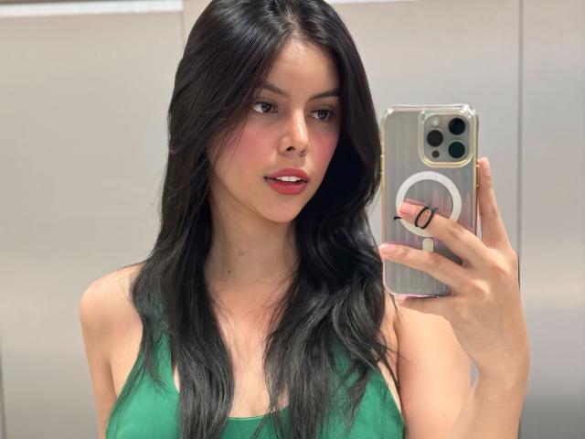 KatC with long black hair, green top, holding phone in right hand, taking mirror selfie, pink lipstick, slight blush, looking at camera, visible phone camera, medium breast size.