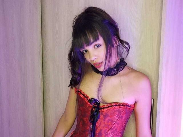 MagicLadies with dark hair, bangs, and purple highlights, wearing a red lace corset with black ribbon, black lace choker, and looking up with a slightly open mouth.