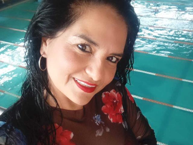 SailorOne has wet black hair, red lipstick, and floral sheer top, smiling with hoop earrings, showing small tits and a cute face, standing by a green-tiled pool.