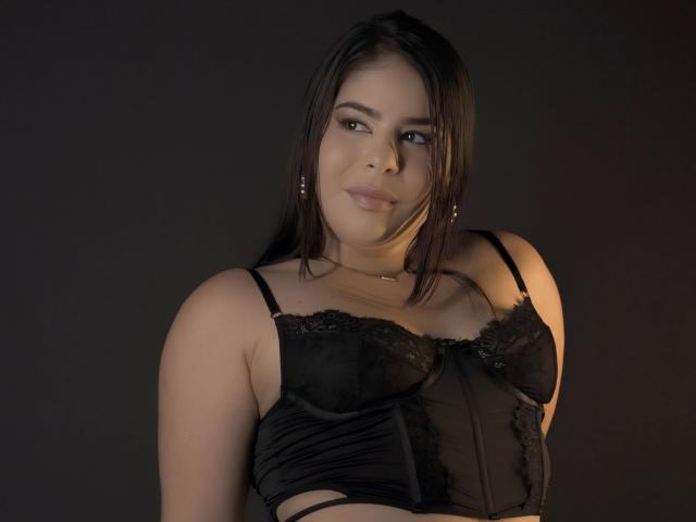 MagicLadies with dark hair, light skin, and small tits in a black lace bra, slightly smiling, wearing a necklace, against a dark background. Her eyes are soft and she's cute.