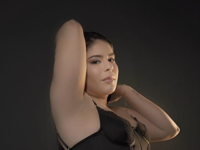 MagicLadies with arms raised, wearing black lace bra, showcasing her tits, dark hair, light skin, subtle makeup, and a confident, sexy expression in a softly lit, dark background.