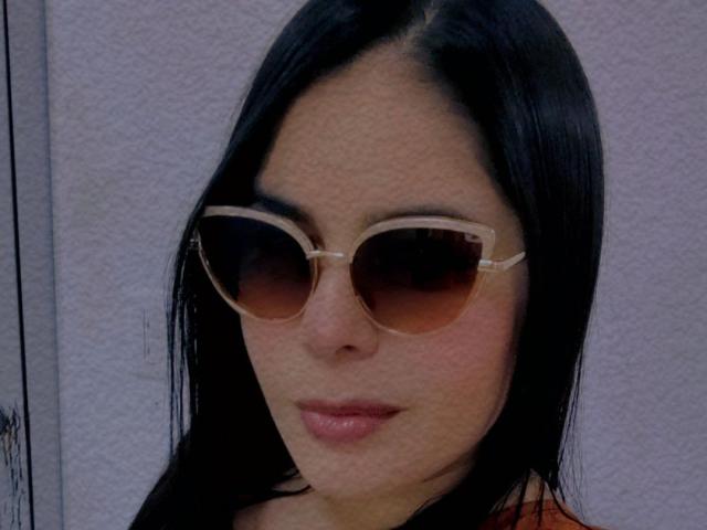 MagicLadies has long black hair, wearing tan cat-eye sunglasses, and a subtle pink lipstick. Her ass is not visible, just her face. She looks slightly bored.