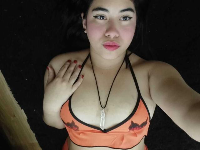 BiancaSolas has orange bikini top with cat prints, black halter, showing her tits, red nails, black necklace, lying on black surface, looking up with pink lips.