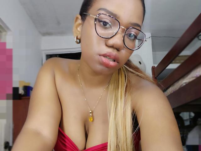 MariieRi with light brown skin, long blonde ponytail, glasses, nose ring, gold necklace, red strapless top, showing her tits, slightly parted lips, in a bedroom.