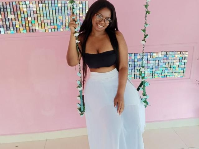 MariieRi with long black hair, glasses, and a smile, wearing a black strapless top and white sheer skirt, sitting on a flower-adorned swing, showing her tits.