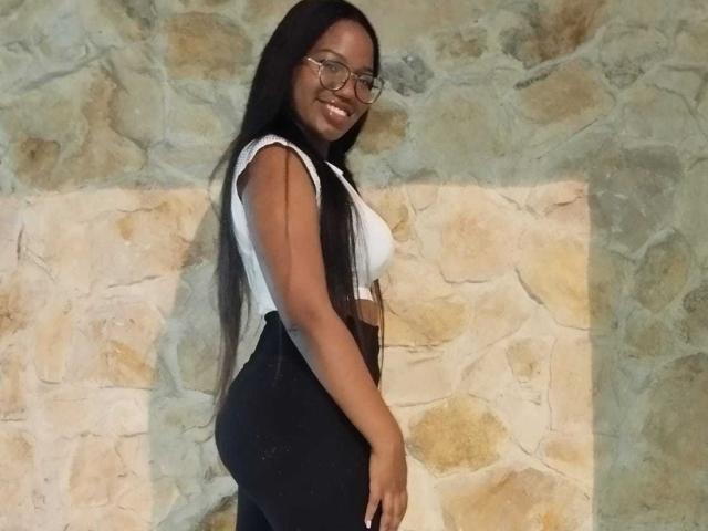 MariieRi with long black hair, glasses, white crop top, and black pants, standing sideways showing her ass and smiling at the camera. Stone wall in the background.