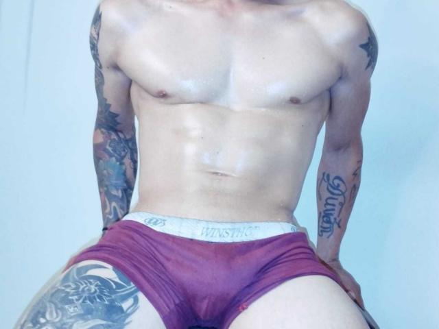 TommyAndGrace, shirtless, with shiny skin, red boxer briefs, tattoos on arms and thigh, sitting, visible abs, muscular, white background, hands on knees, visible chest and torso.