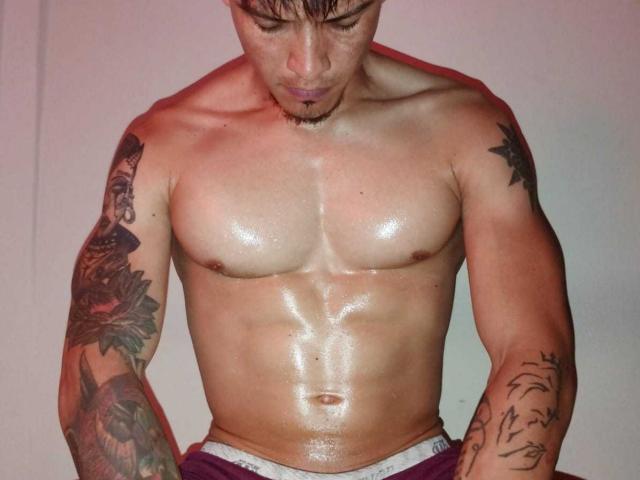 TommyAndGrace, shirtless, glistening with sweat, muscular, tattooed arms, looking down, dark hair, light skin, slightly shiny abs, wearing purple pants with white waistband, red lighting.