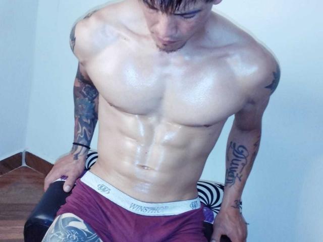 TommyAndGrace, torse nu avec des abdominaux brillants et huilés et des tatouages, portant des sous-vêtements bordeaux, assis sur une chaise avec un coussin rayé, regardant vers le bas avec des cheveux bruns courts..