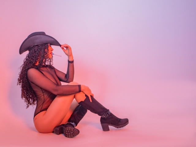 MilahLove sits on the floor, wearing a black mesh bodysuit, cowboy hat, and boots. She's holding glasses, with curly hair and a sexy, confident vibe.