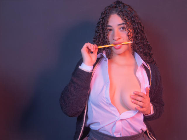 MilahLove with curly brown hair, black cardigan, and unbuttoned white shirt, holding a pencil in her mouth, exposing her large tits with one hand covering her nipple.