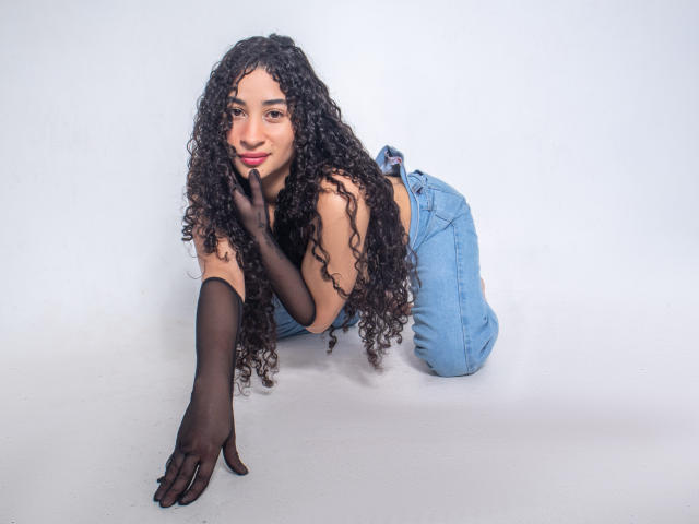 MilahLove with curly black hair, wearing black sheer glove, blue jeans, and red lipstick, on her knees, hand on floor, smiling, showing off her tits and cute ass.