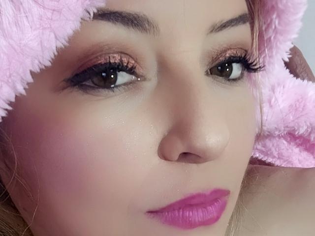NatalyHami's face, wearing a pink fuzzy hat, with pink lipstick, brown eyes, and subtle makeup, showing a gentle, slightly curious expression. Her skin is smooth, with a slight blush.